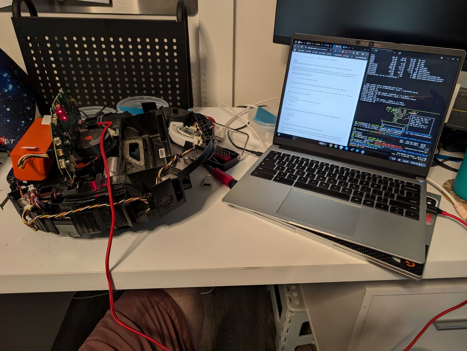 A disassembled robotic vacuum cleaner lying on a desk. There is a laptop computer also on the desk, with code visible on the screen. Several cables and a screwdriver are also visible on the desk. The screen shows code related to the robot vacuum.